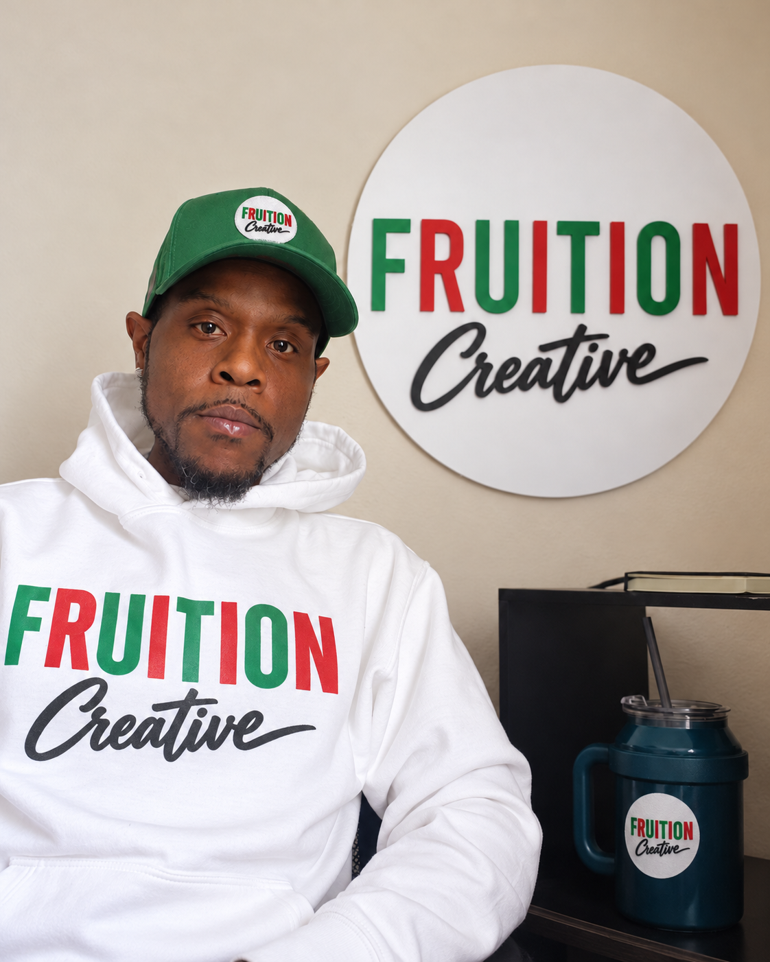 Bobby Smith beside the Fruition Creative sign, with coordinated Fruition Creative branding on his hoodie, cap and cup